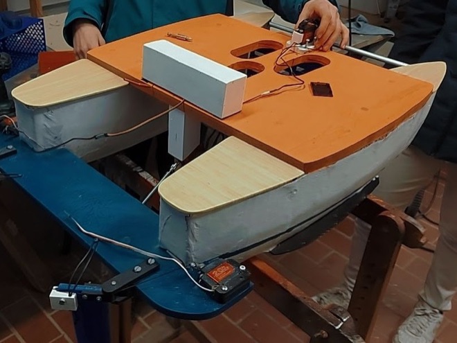 A radio-controlled trimaran part-way through a build on a workbench, with a wooden deck, foam hulls, and a servo wired to the rudder linkage.