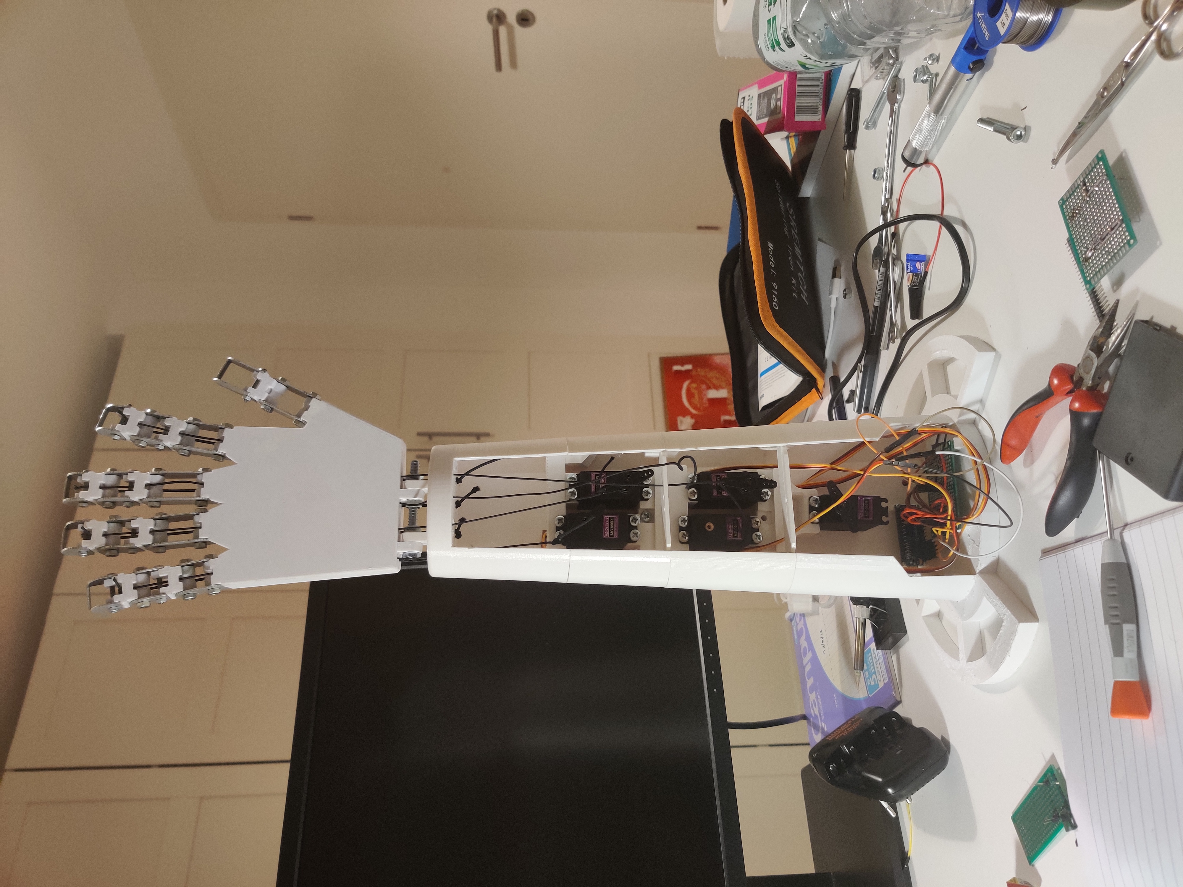 A white 3D-printed robotic arm and articulated gripper hand, wired to a row of servos, on a workbench.